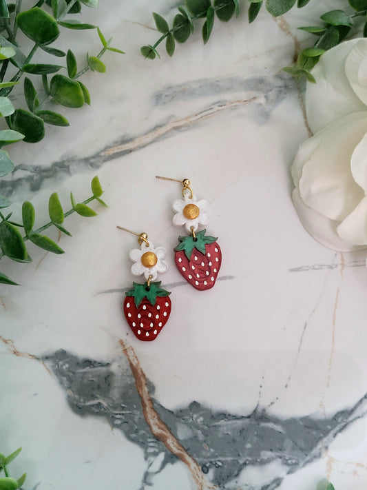 Close up of Resin Strawberry and flower earrings with gold findings on a marble background surrounded by foliage.