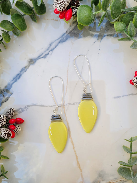 close up of yellow christmas lightbulb earrings on a marble background surrounded by foliage.