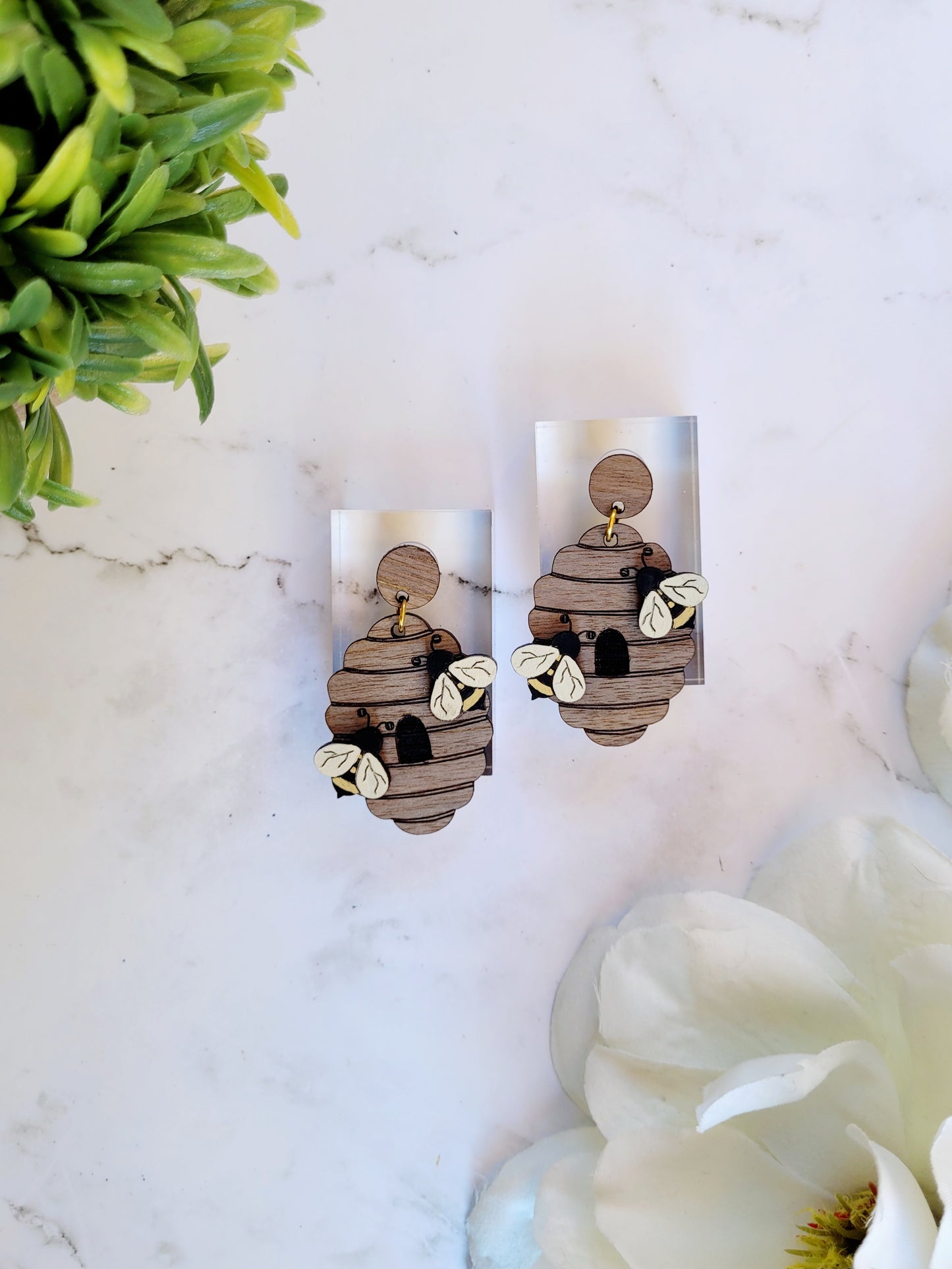 wooden beehive shaped earrings with bee charms on a marble background surrounded by foliage.