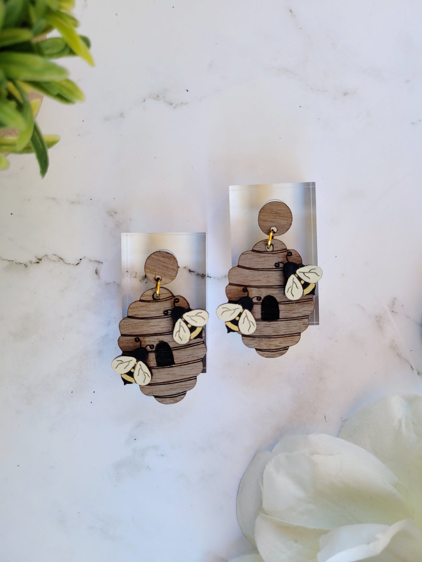 close up of wooden beehive shaped earrings with bee charms on a marble background surrounded by foliage.