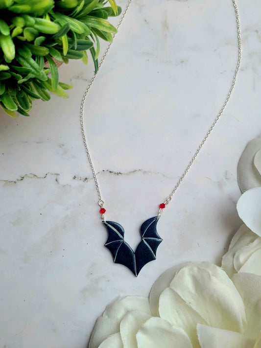 close up of red bat boy necklaces on a marble background surrounded by foliage.