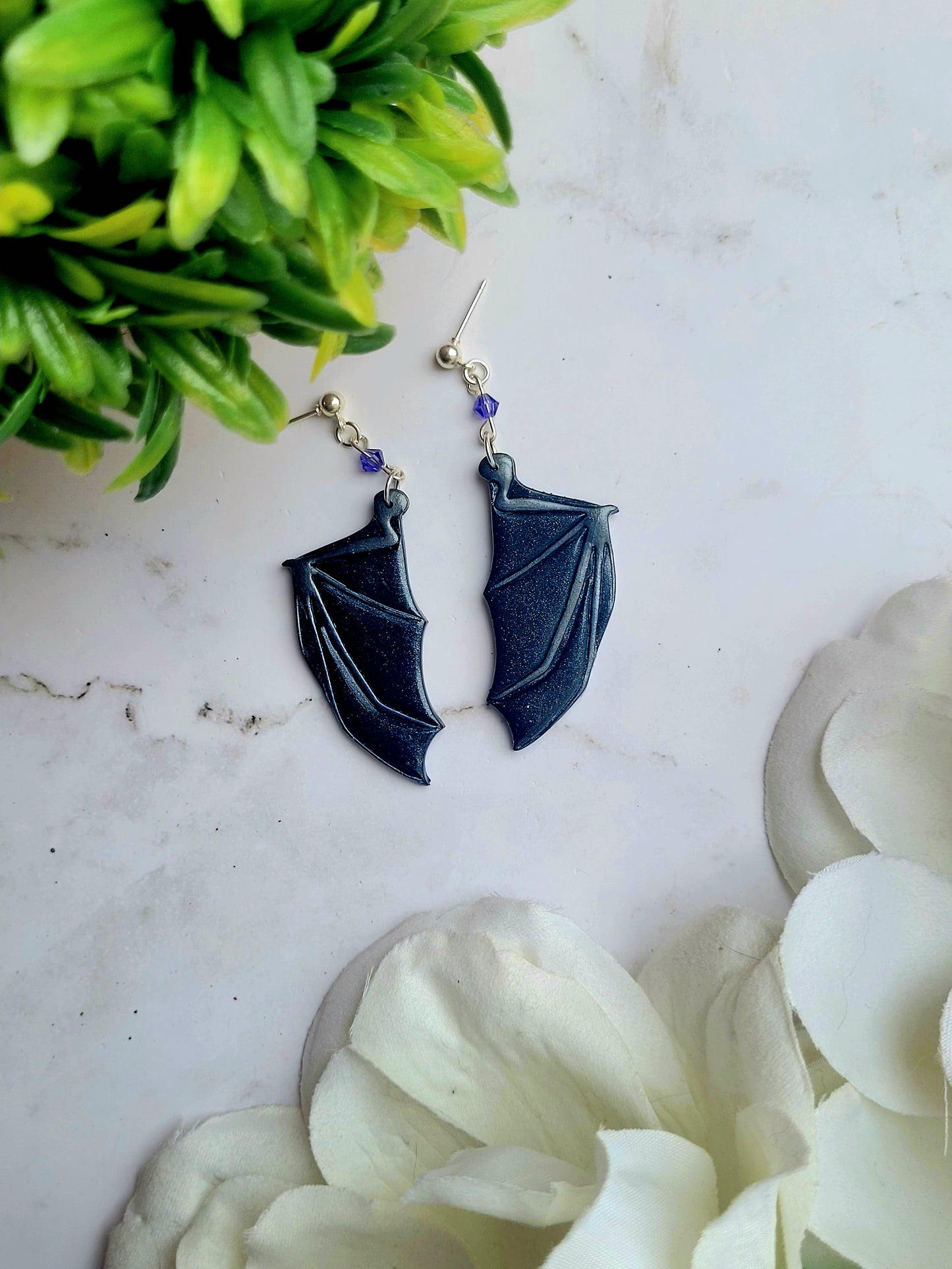close up of purple bat boy earrings on a marble background surrounded by foliage.