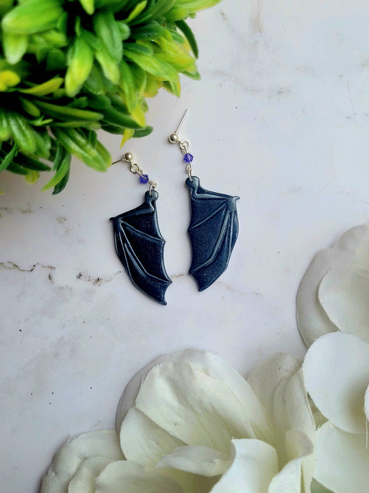 close up of purple bat boy earrings on a marble background surrounded by foliage.