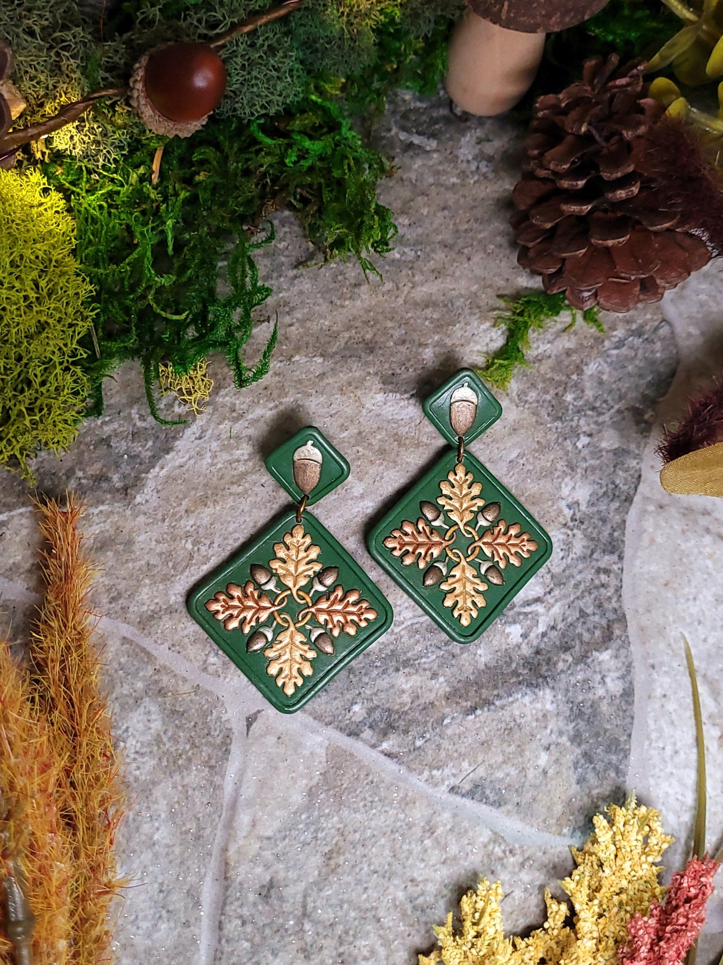Green diamond shaped earrings with metallic acorns and oak leaves. Sitting on a stone background surrounded by foliage.