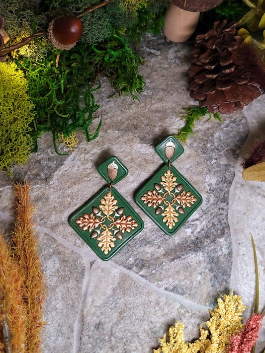 Green diamond shaped earrings with metallic acorns and oak leaves. Sitting on a stone background surrounded by foliage.