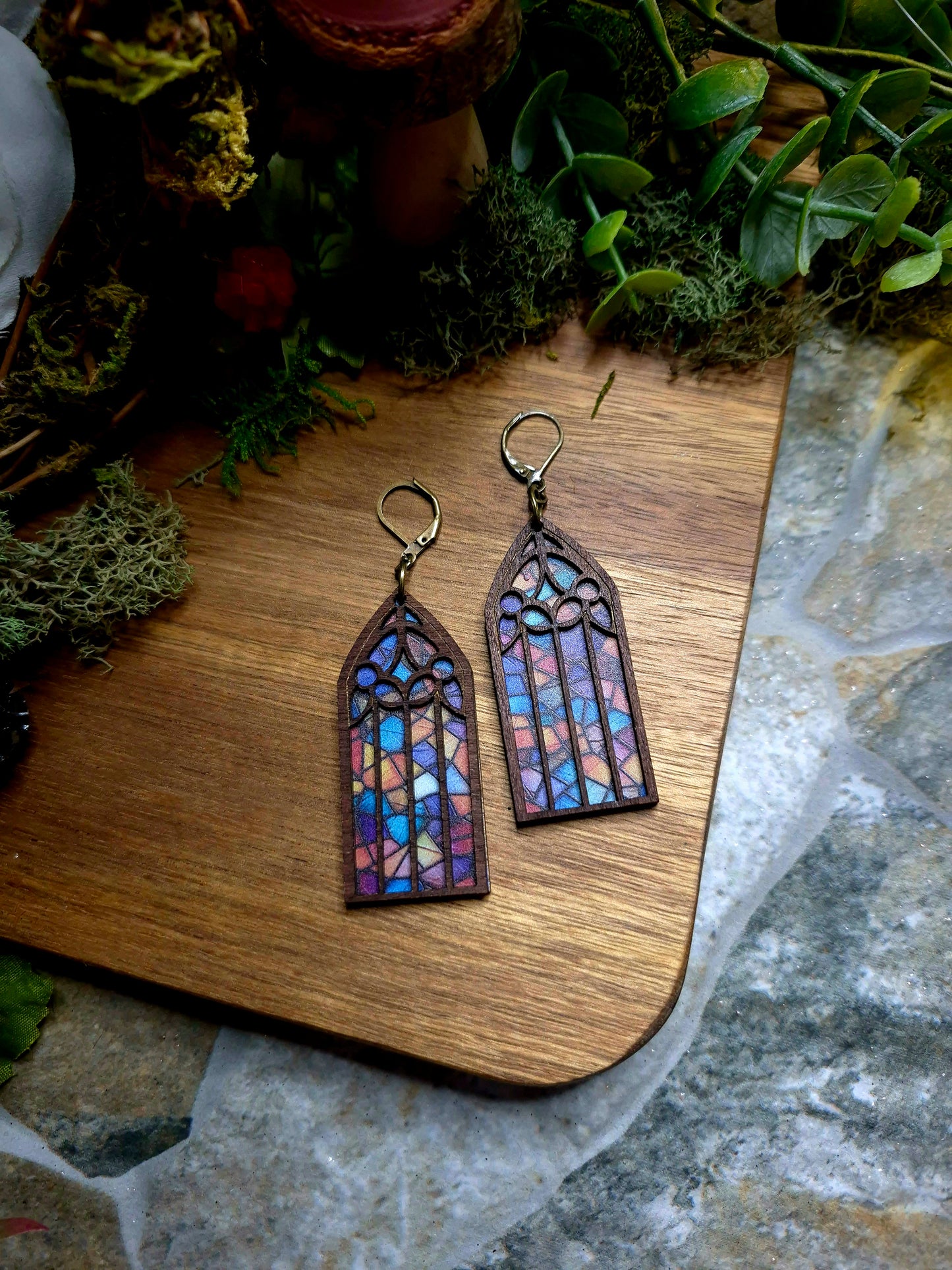 Rainbow Cathedral window earrings on a stone background surrounded by foliage.