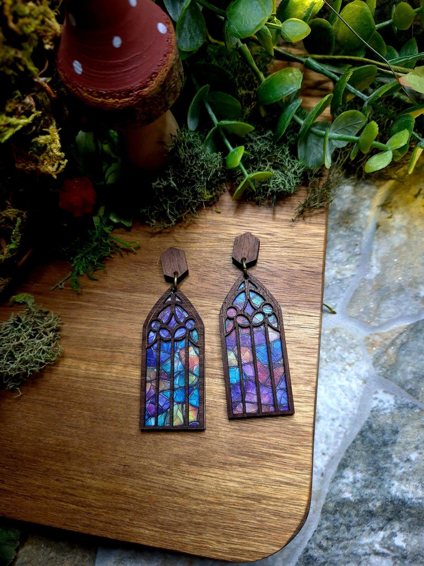 Jewel Cathedral window earrings on a wood and stone background surrounded by foliage.