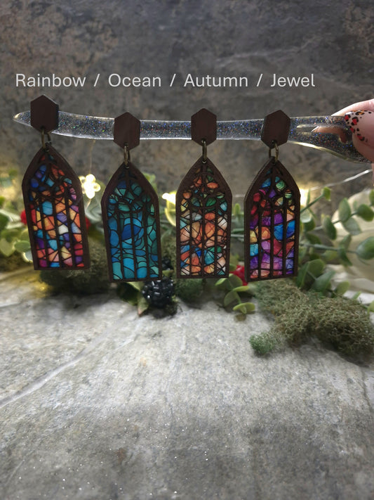 4 Colors of Cathedral window earrings with light shining through on a stone background surrounded by foliage.