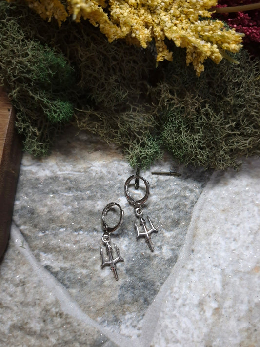 Silver trident earrings on a stone background surrounded by foliage