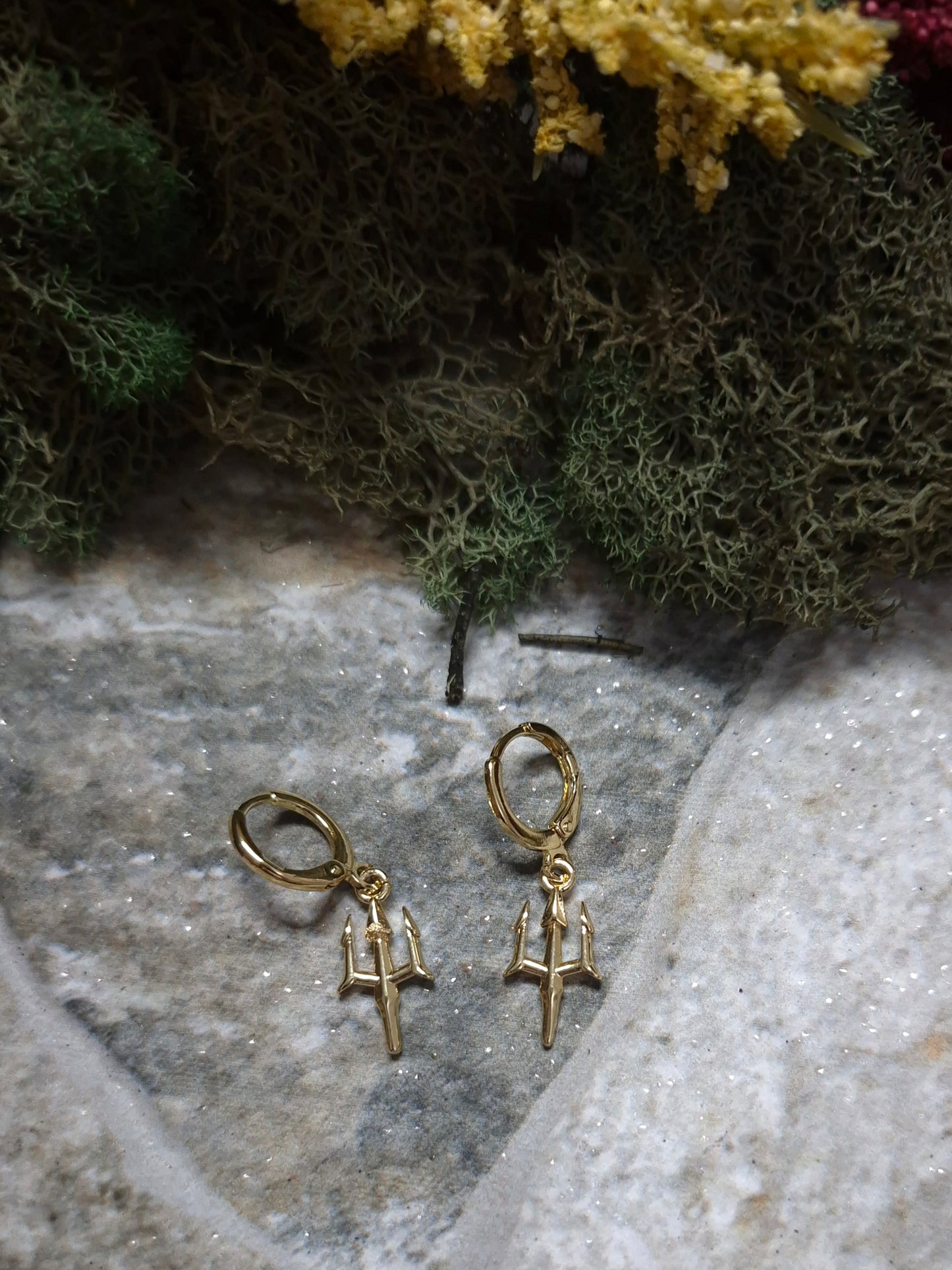 Gold trident earrings on a stone background surrounded by foliage