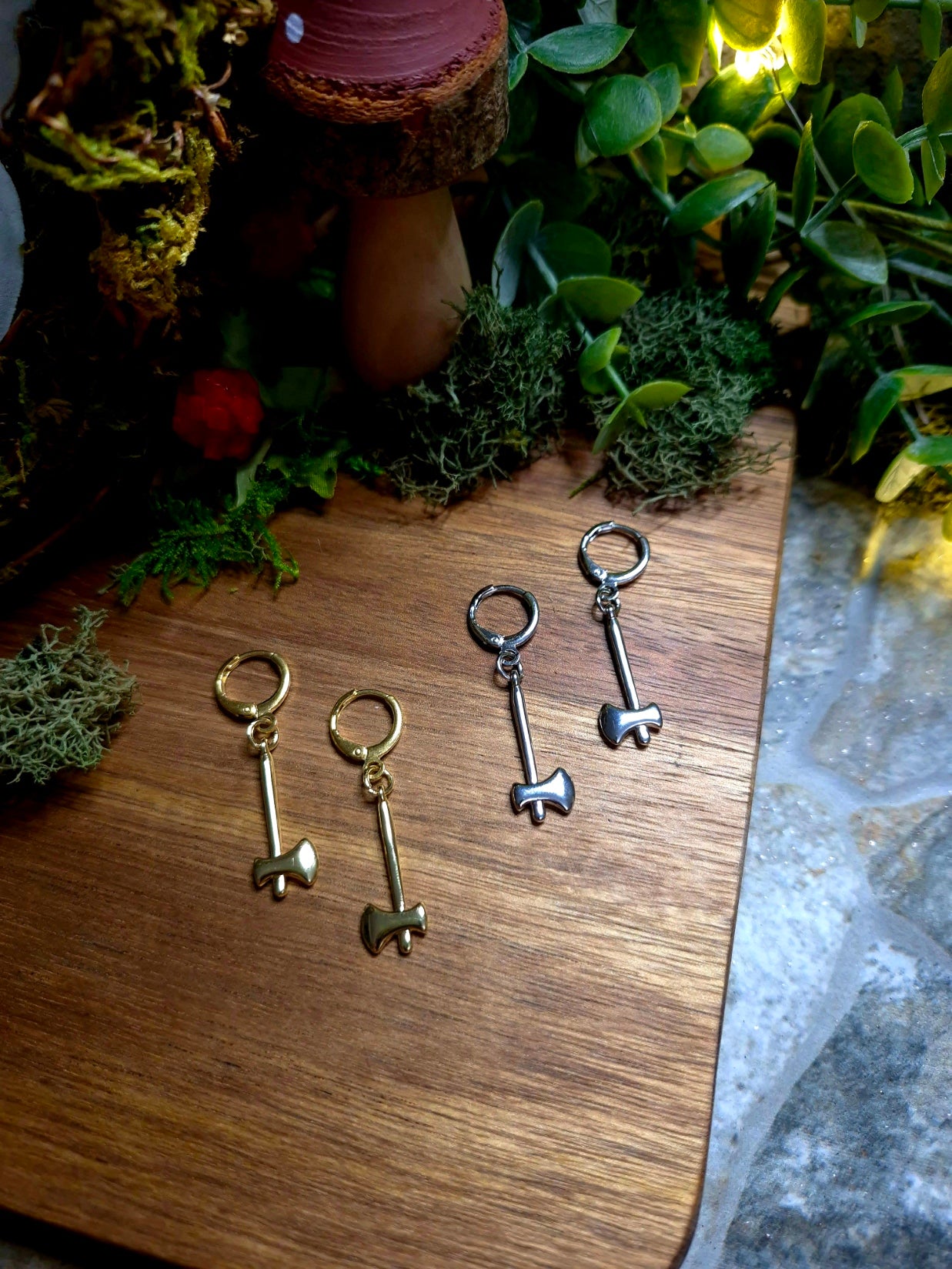 Gold and Silver Axe Earrings on a wood and stone background surrounded by foliage
