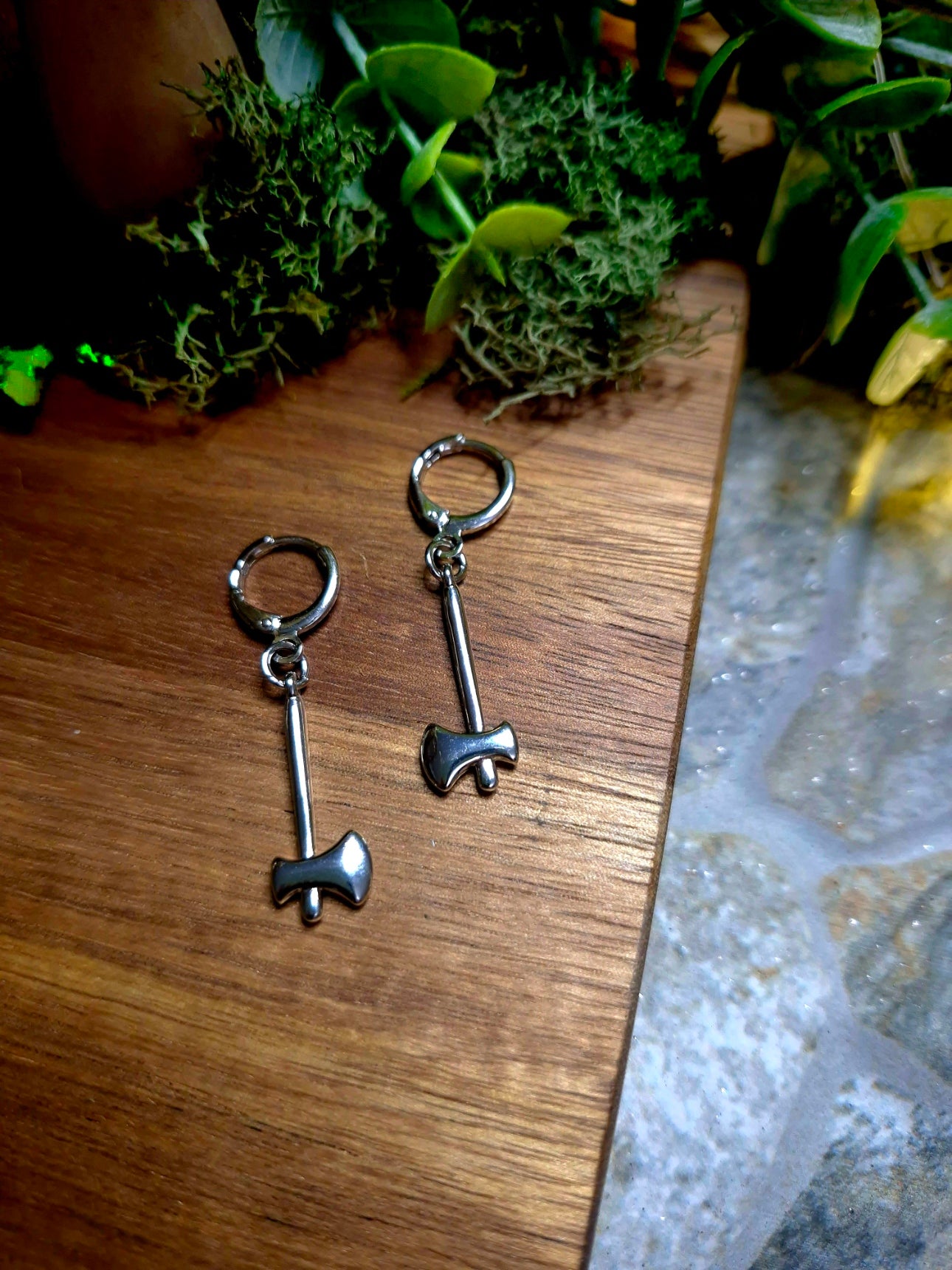 Silver Axe Earrings on a wood and stone background surrounded by foliage