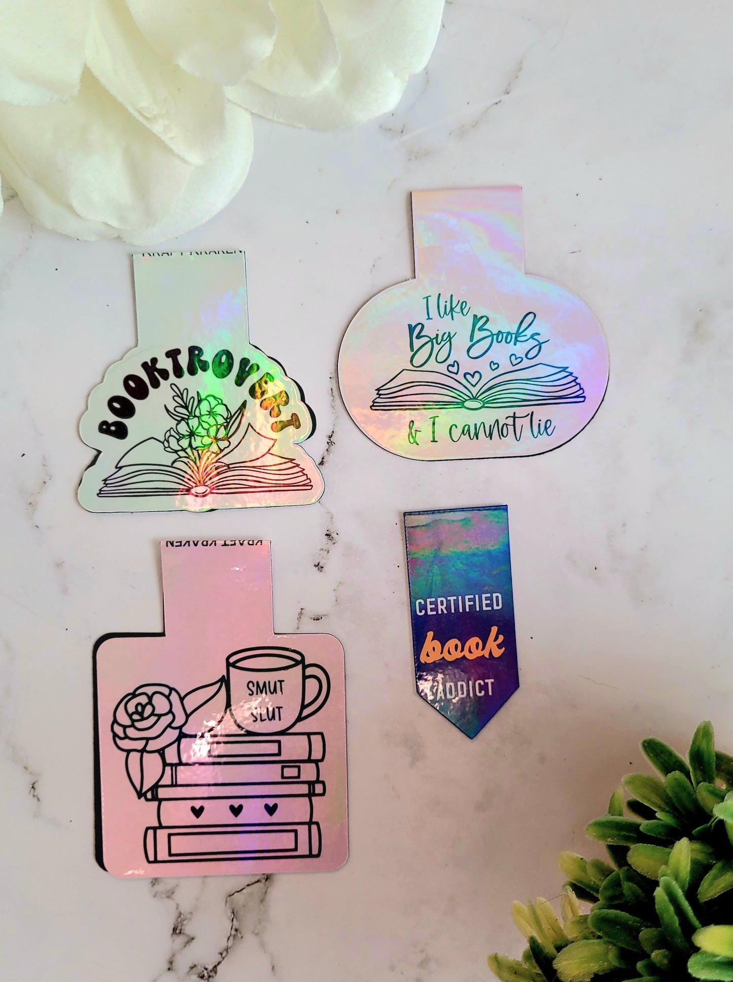 Booktrovert, book addict, big books, smut slut bookmarks on a marble background surrounded by foliage.