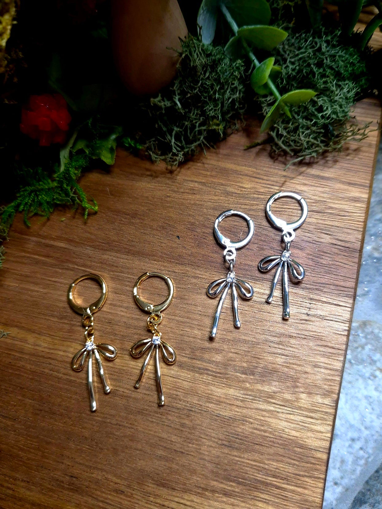 Silver and Gold Bow earrings on a wood and stone background surrounded by foliage