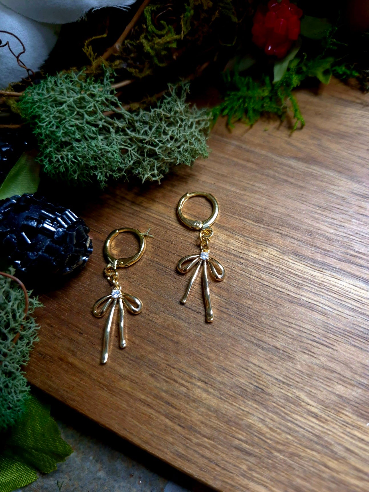 Gold Bow earrings on a wood and stone background surrounded by foliage