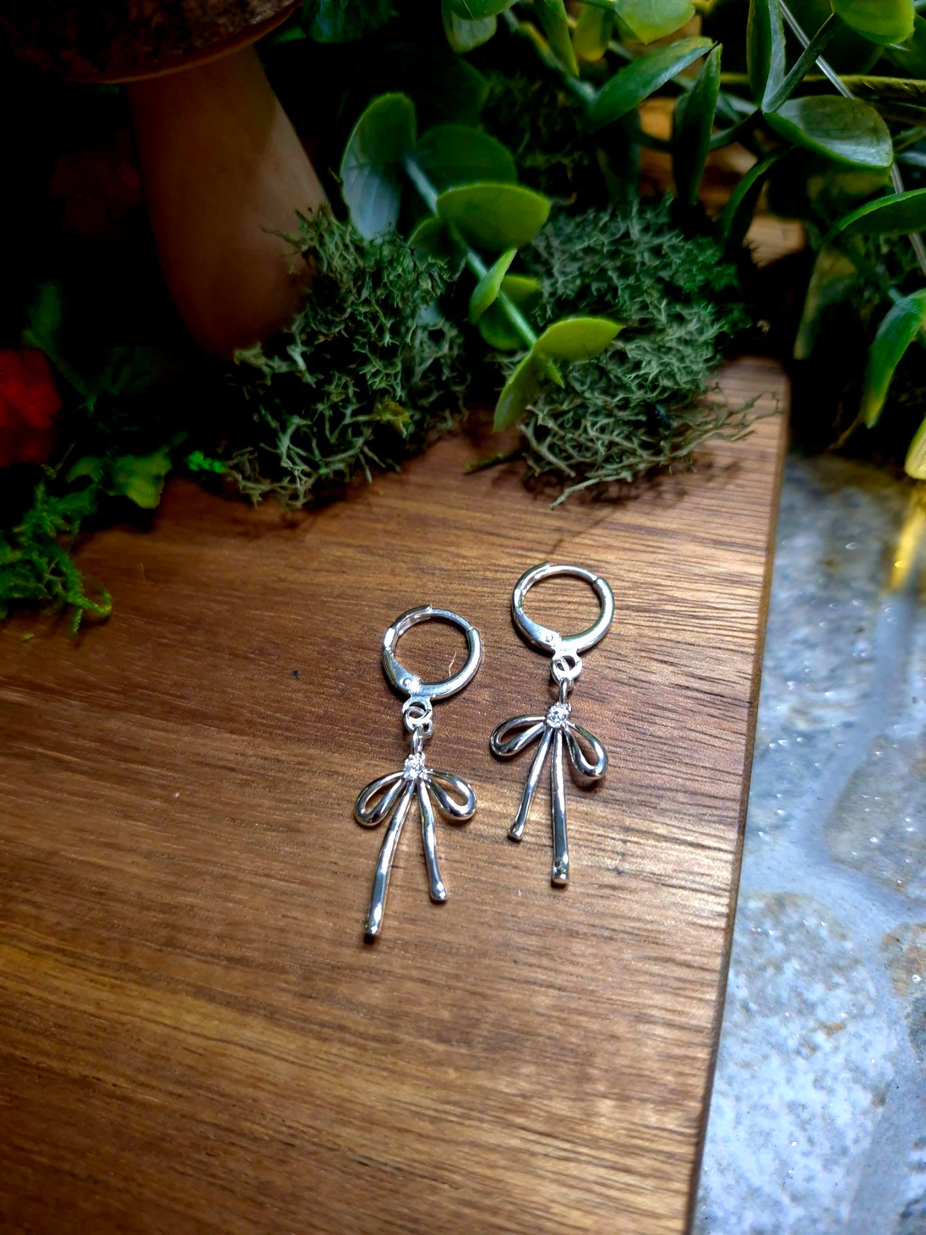 Silver Bow earrings on a wood and stone background surrounded by foliage