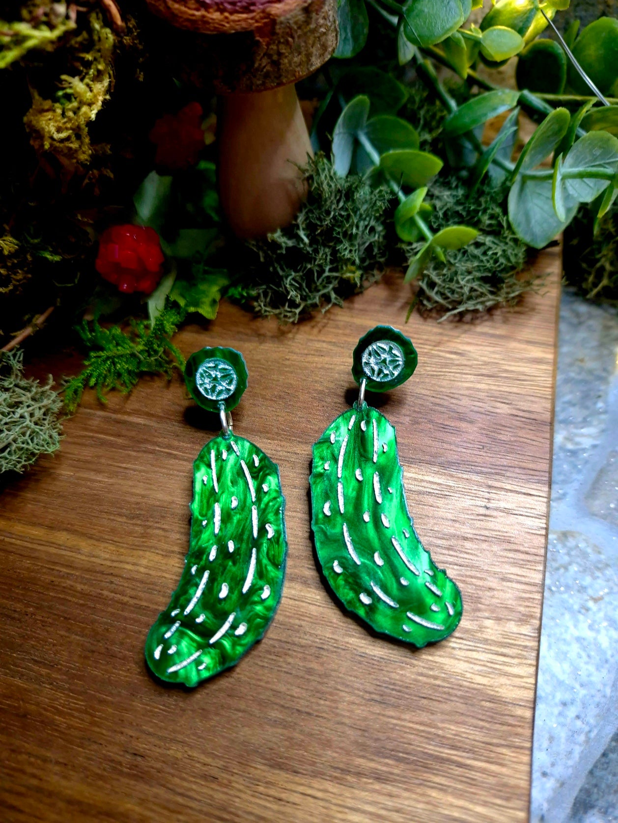 Green pickle earrings on a wood and stone background surrounded by foliage.