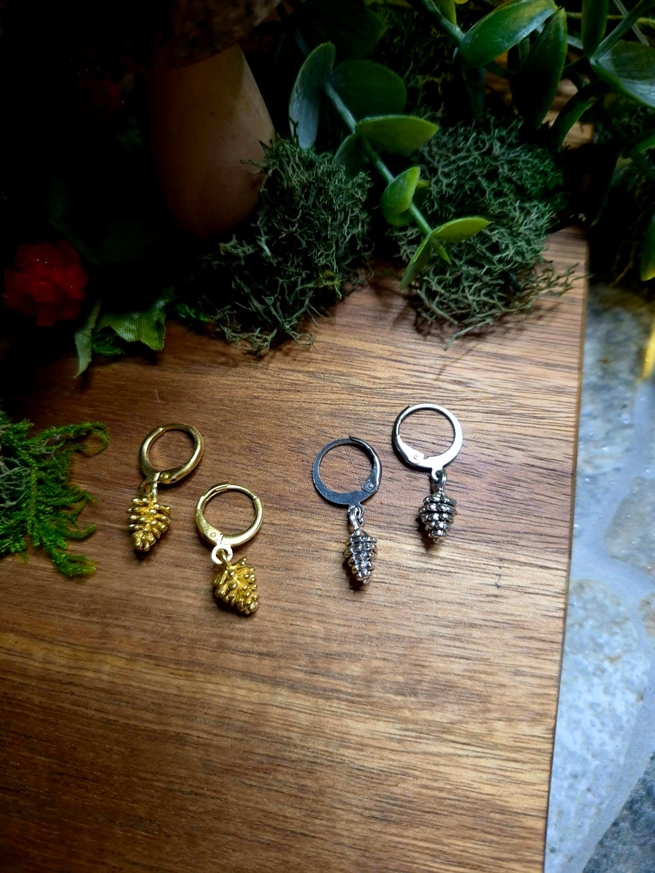 silver and gold pinecone earrings on a wood and stone background surrounded by foliage.