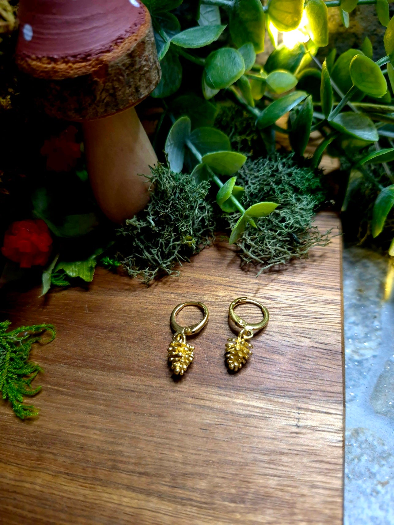 gold pinecone earrings on a wood and stone background surrounded by foliage.