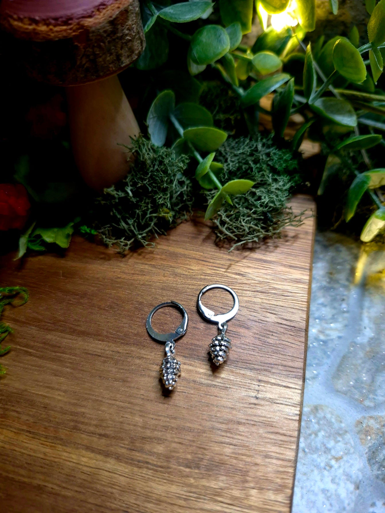 silver pinecone earrings on a wood and stone background surrounded by foliage.