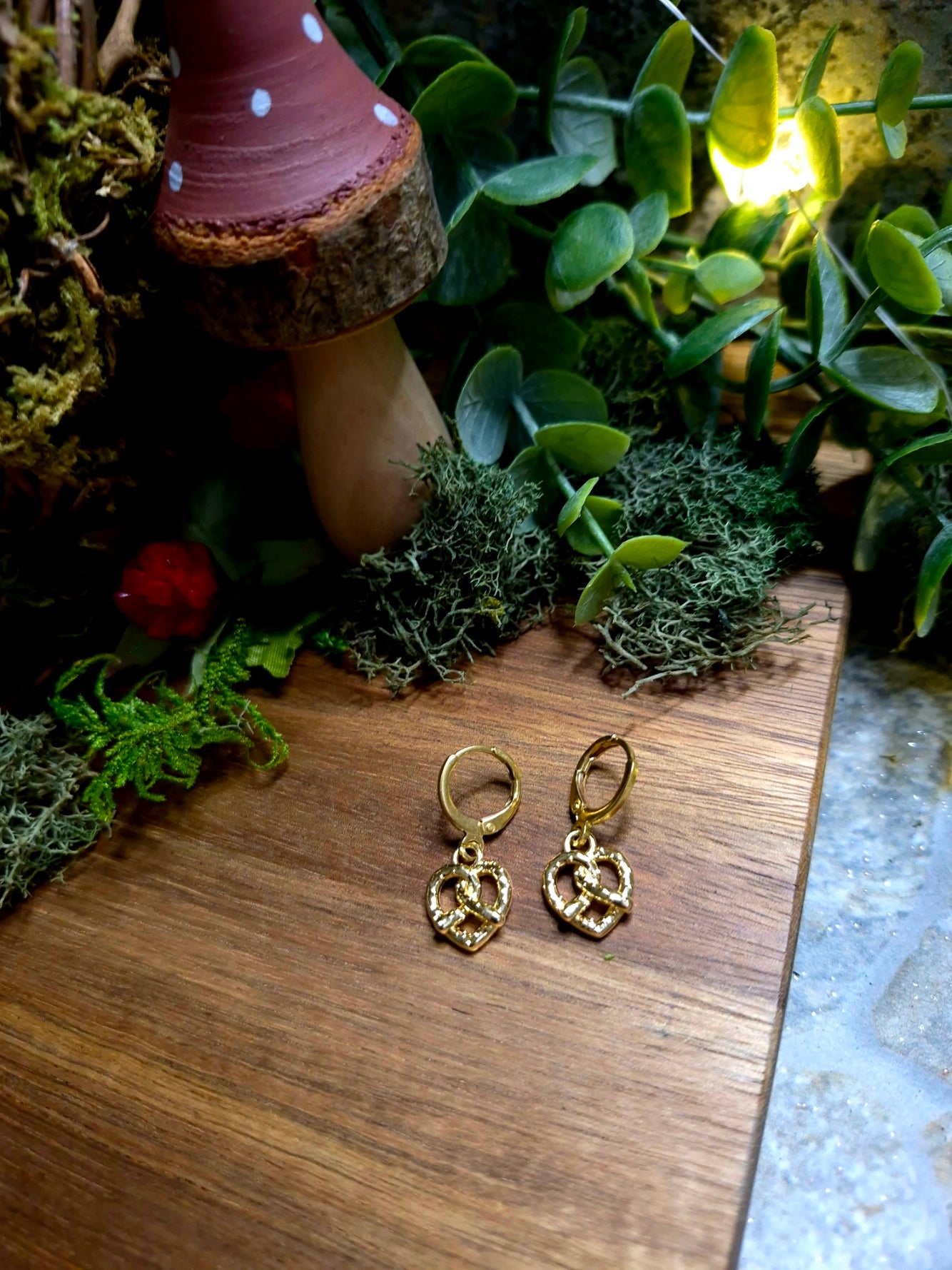 gold pretzel earrings on a wood and stone background surrounded by foliage.