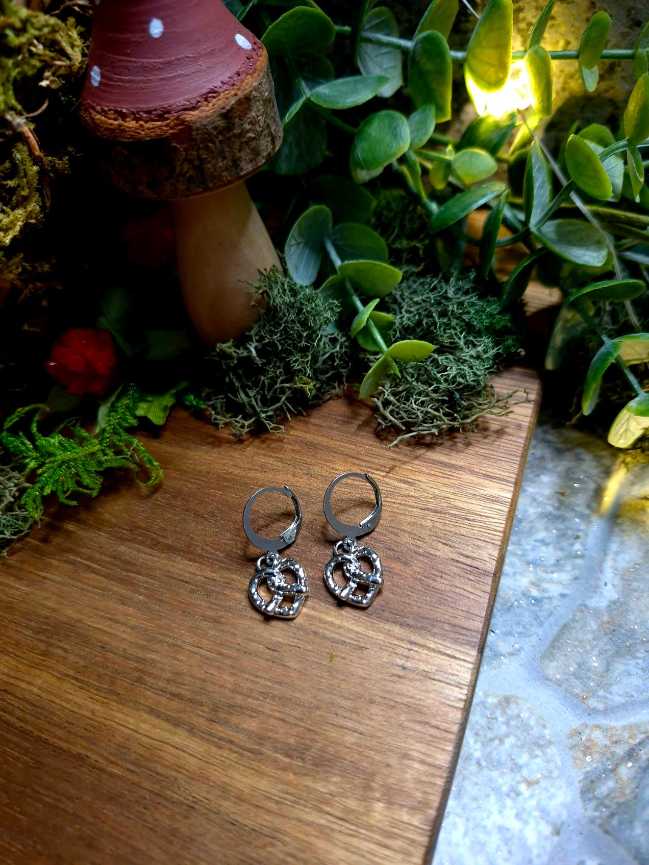 silver pretzel earrings on a wood and stone background surrounded by foliage.