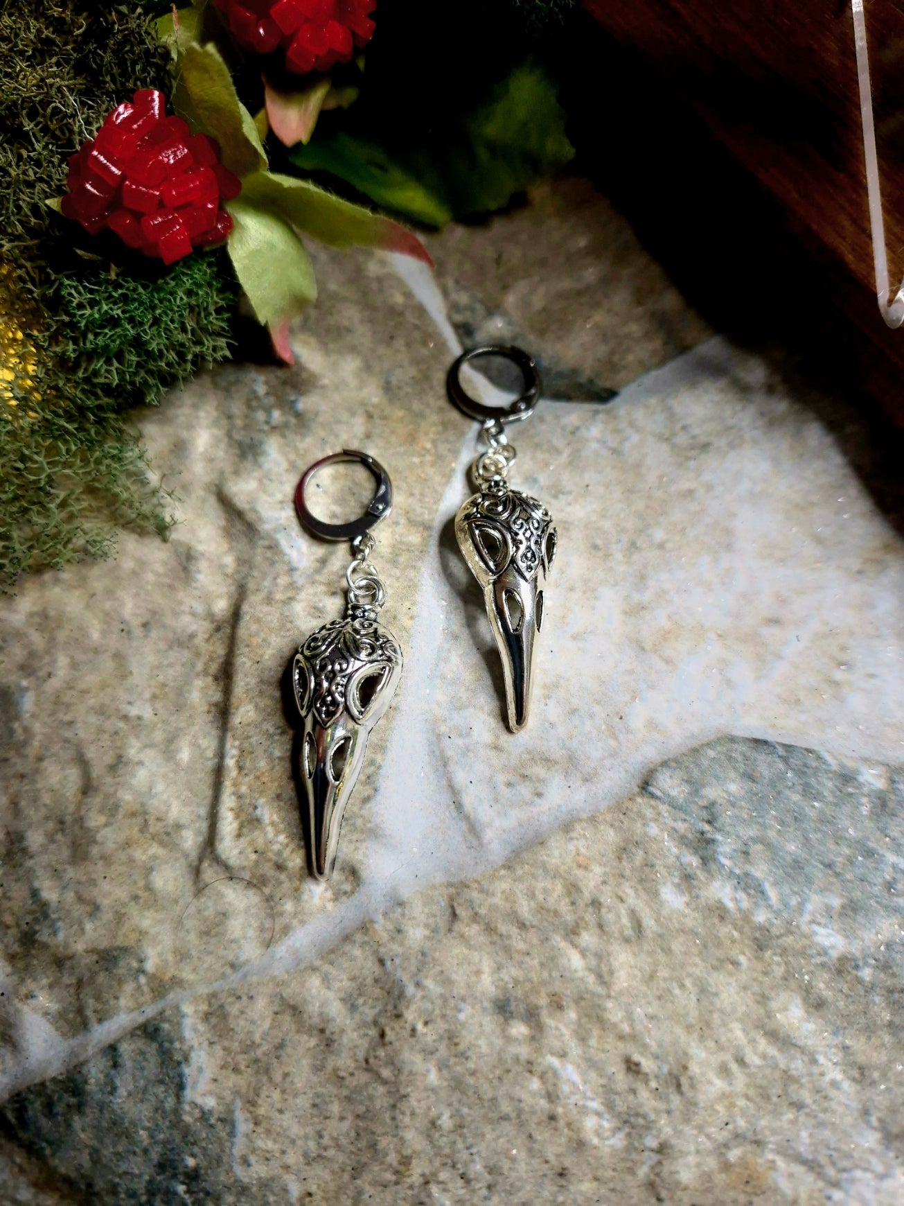 silver raven skull earrings on a stone background surrounded by foliage.