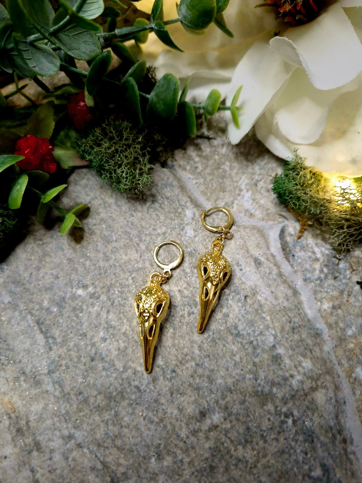gold raven skull earrings on a stone background surrounded by foliage.