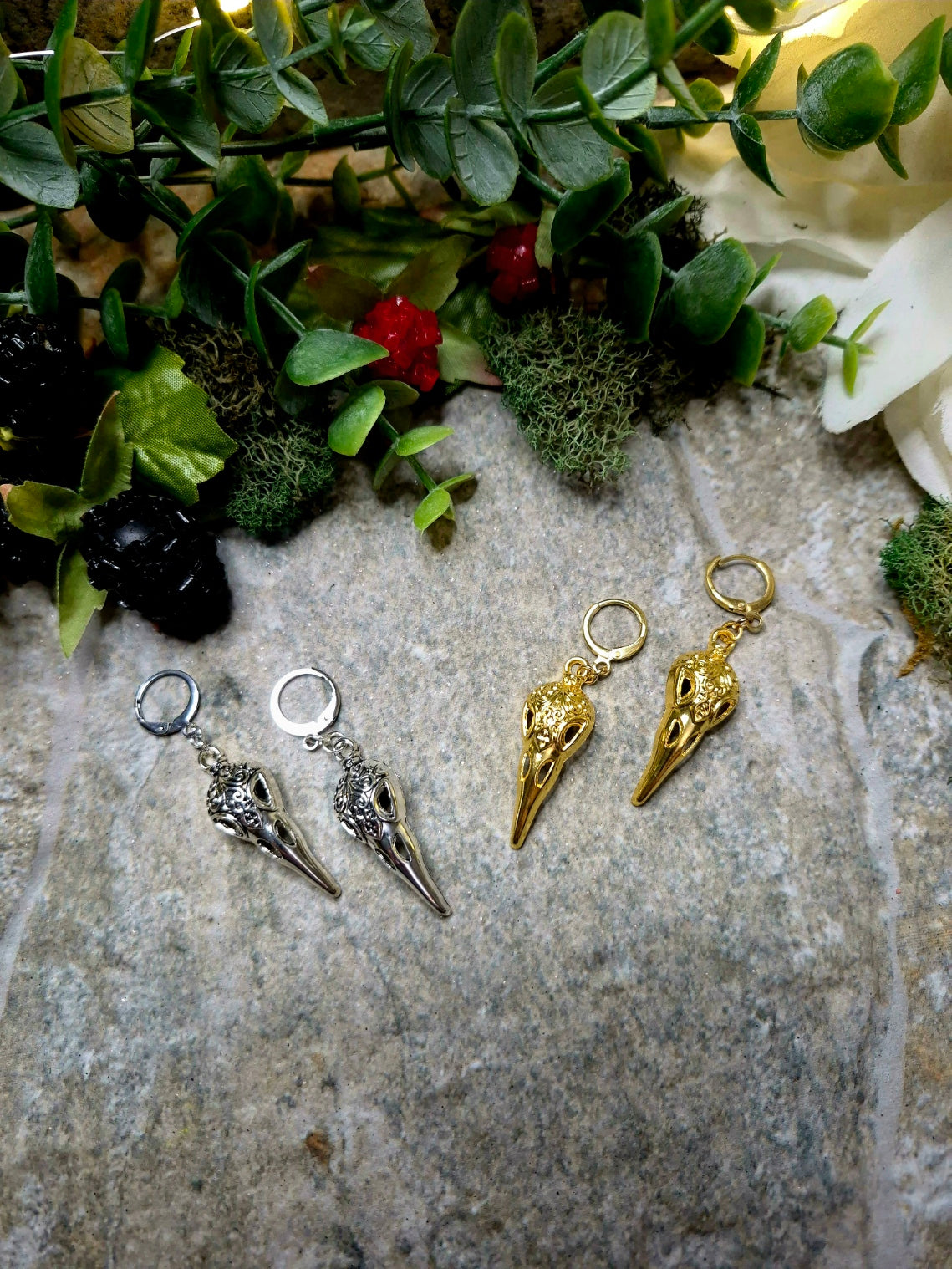 silver and gold raven skull earrings on a stone background surrounded by foliage.