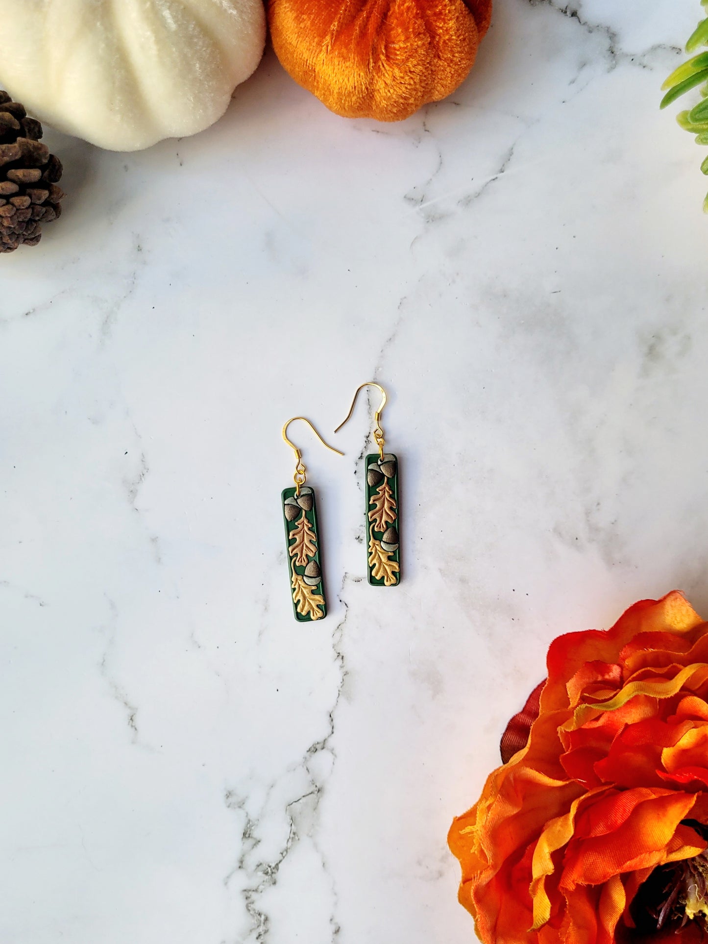 Green bar shaped earrings with metallic acorns and oak leaves. The earrings are on a white marble background with pumpkins and flowers in the background.