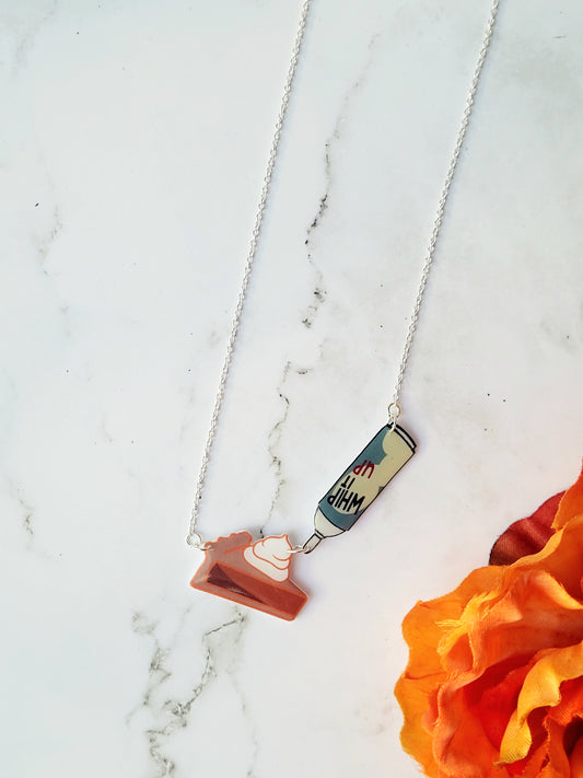 close up of Pumpkin pie slice necklace with a can of whipped cream on a marble background surrounded by fall foliage.