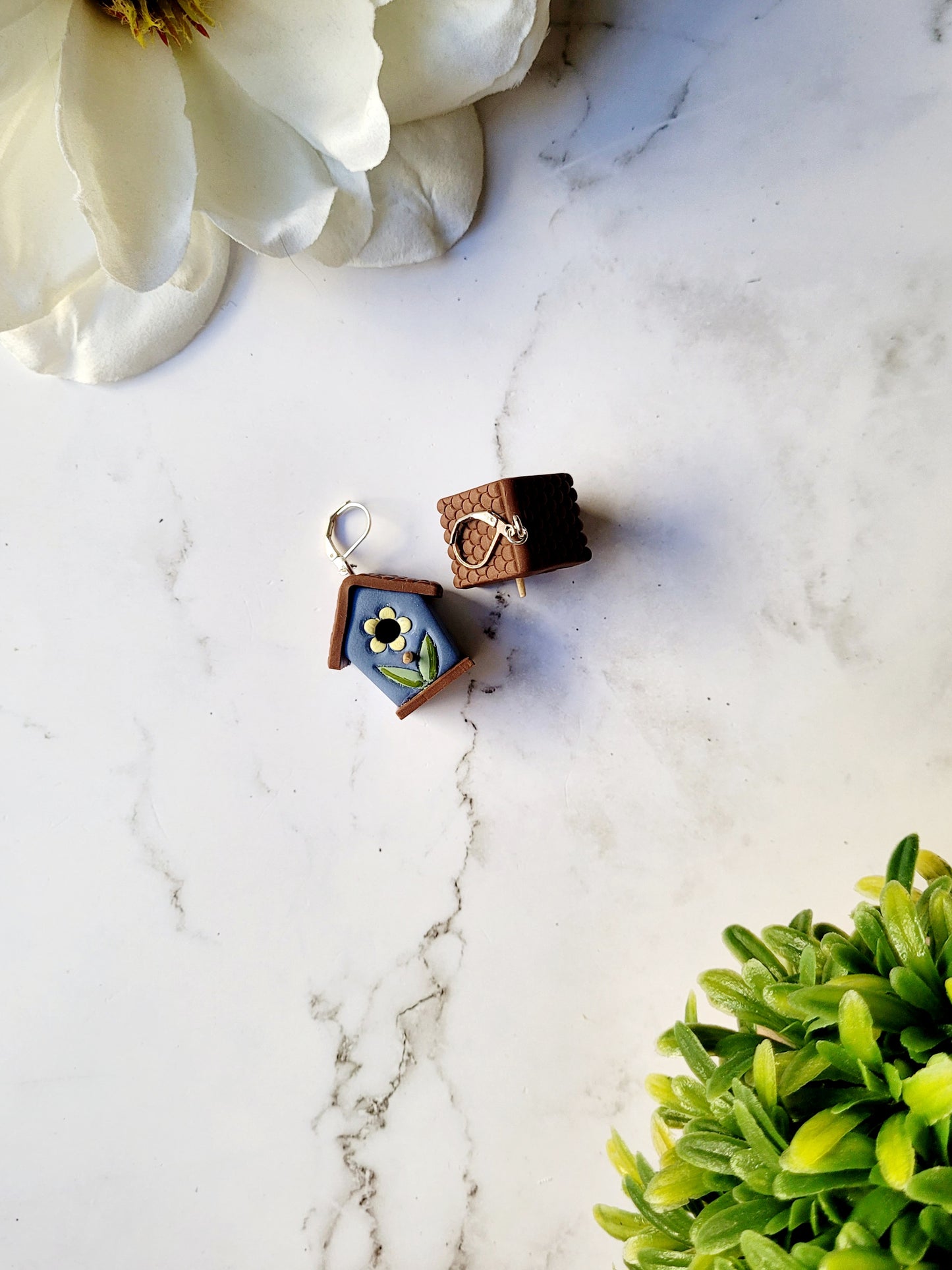 3-d blue birdhouse earrings on a white marble background surrounded by foliage.