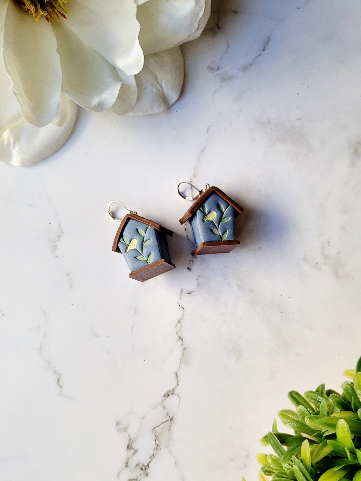 the back of the 3-d blue birdhouse earrings on a white marble background surrounded by foliage.