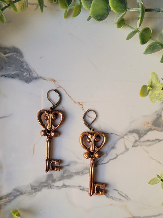 close up of heart skeleton key earrings with filigree in antique gold on a marble background with foliage.