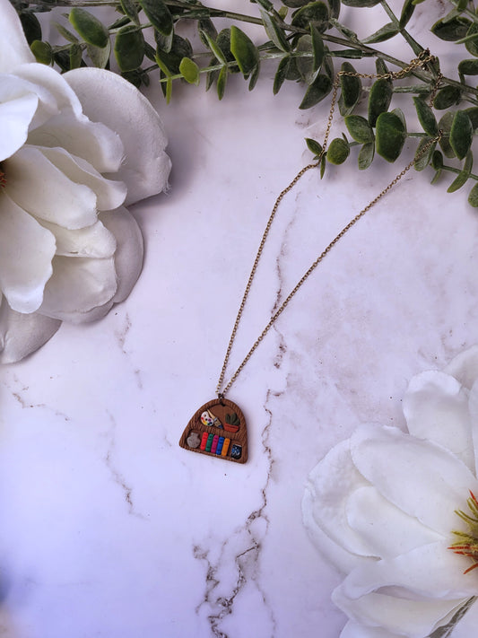 Bookshelf pendant made of polymer clay sits on a marble background surrounded by foliage. 