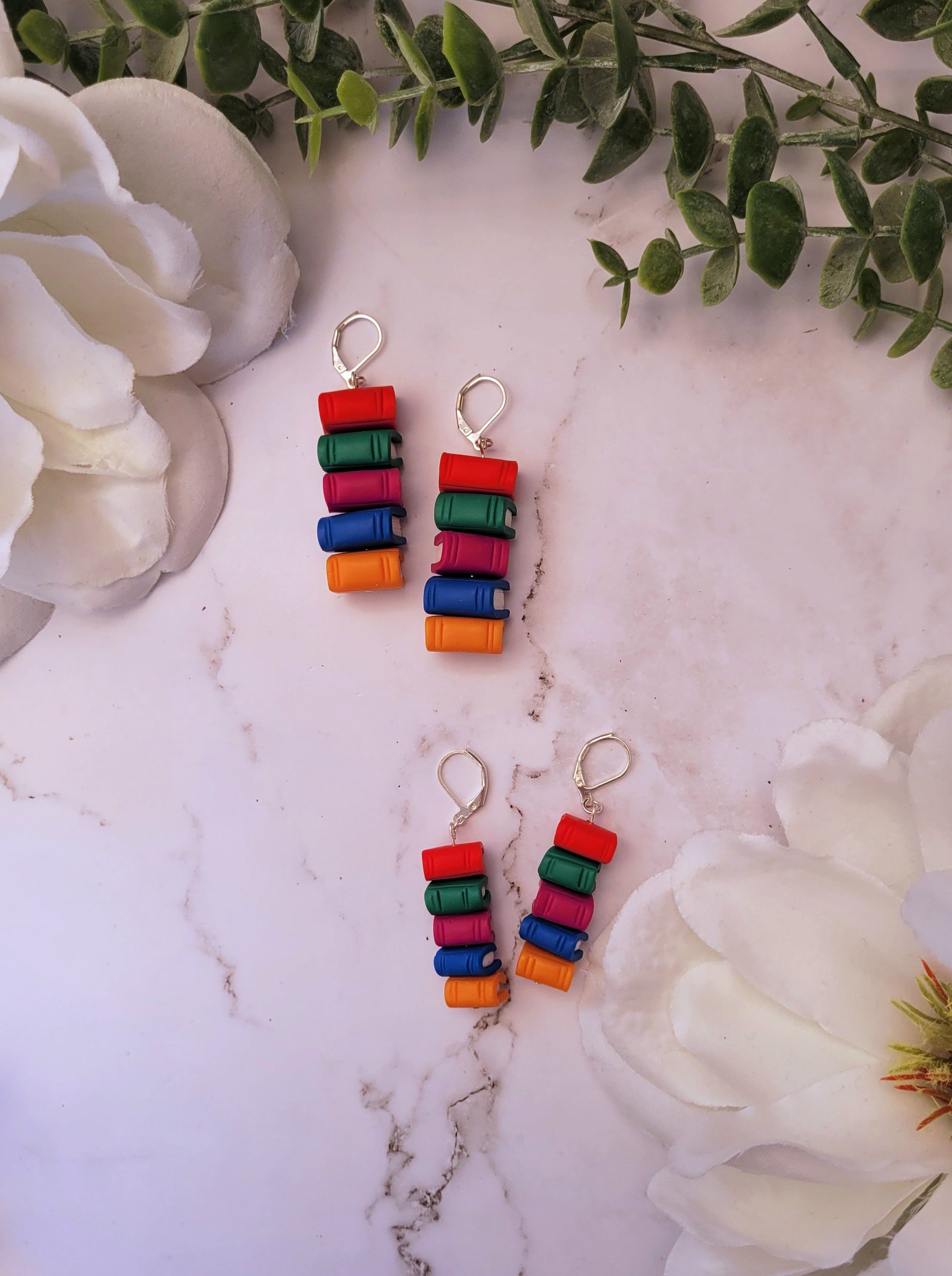 Bookstack earrings made of polymer clay in two sizes sits on a marble background surrounded by foliage.