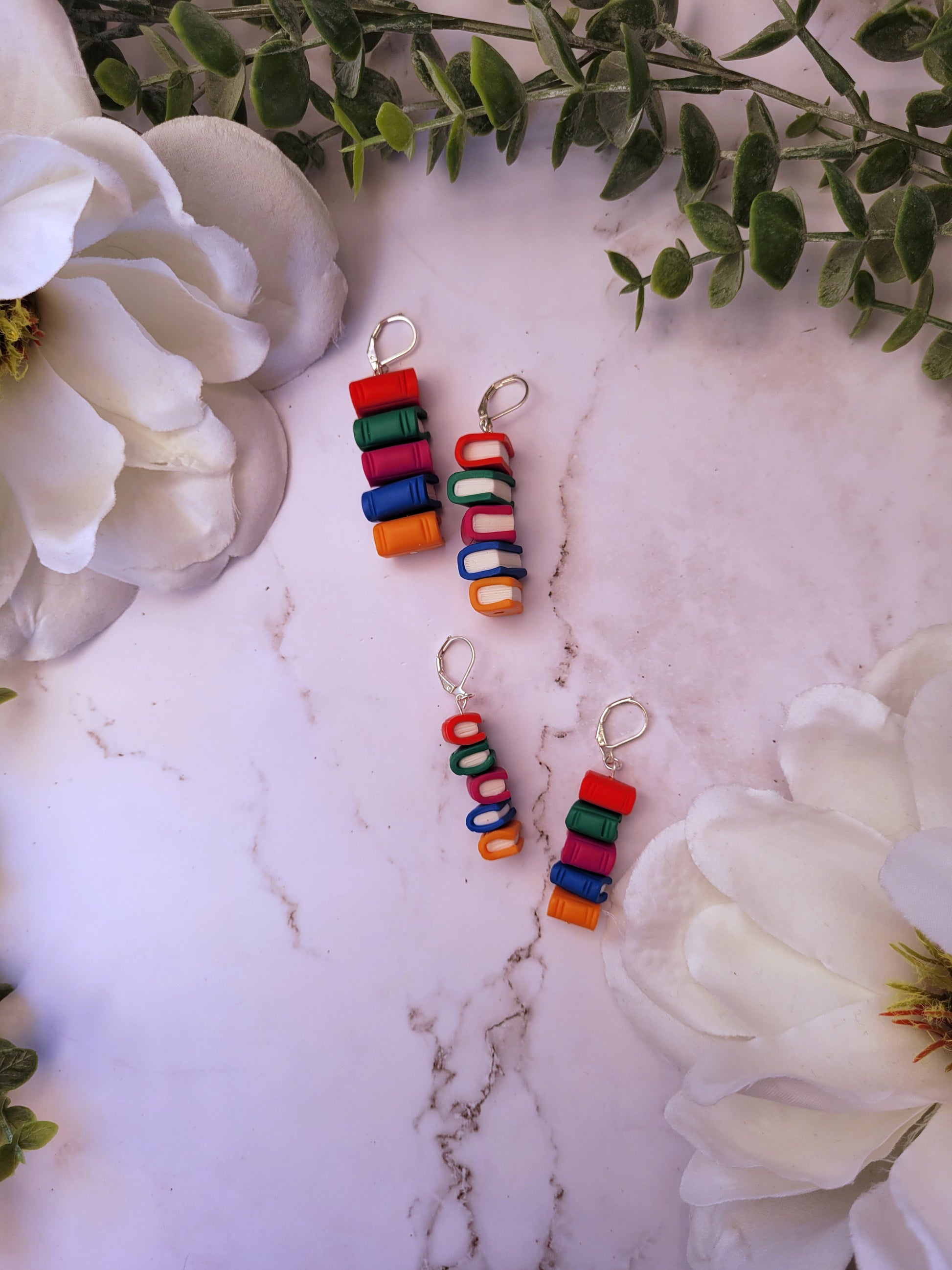 Side view of bookstack earrings made of polymer clay in two sizes sits on a marble background surrounded by foliage.