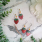 Resin Strawberry and flower earrings with gold and silver findings on a marble background surrounded by foliage. 