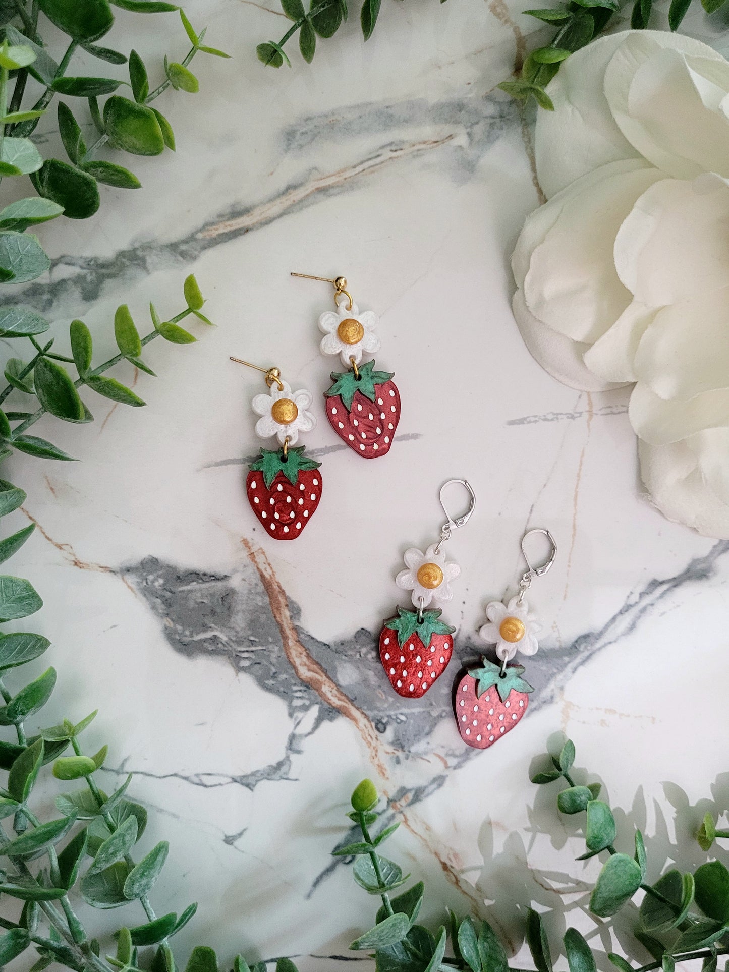 Resin Strawberry and flower earrings with gold and silver findings on a marble background surrounded by foliage. 