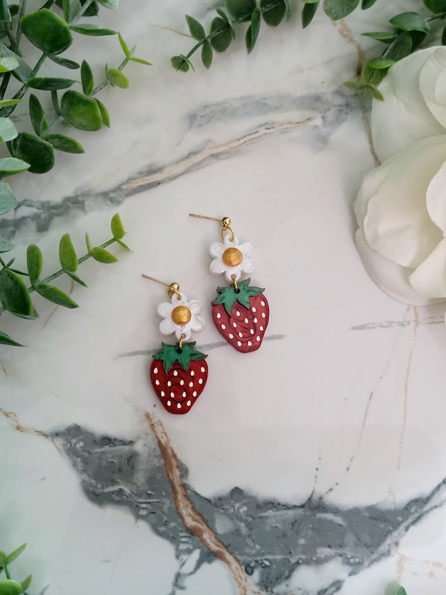 Close up of Resin Strawberry and flower earrings with gold findings on a marble background surrounded by foliage.