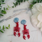 close up of Red Lobster with a blue shell stud earrings on a marble background with foliage.