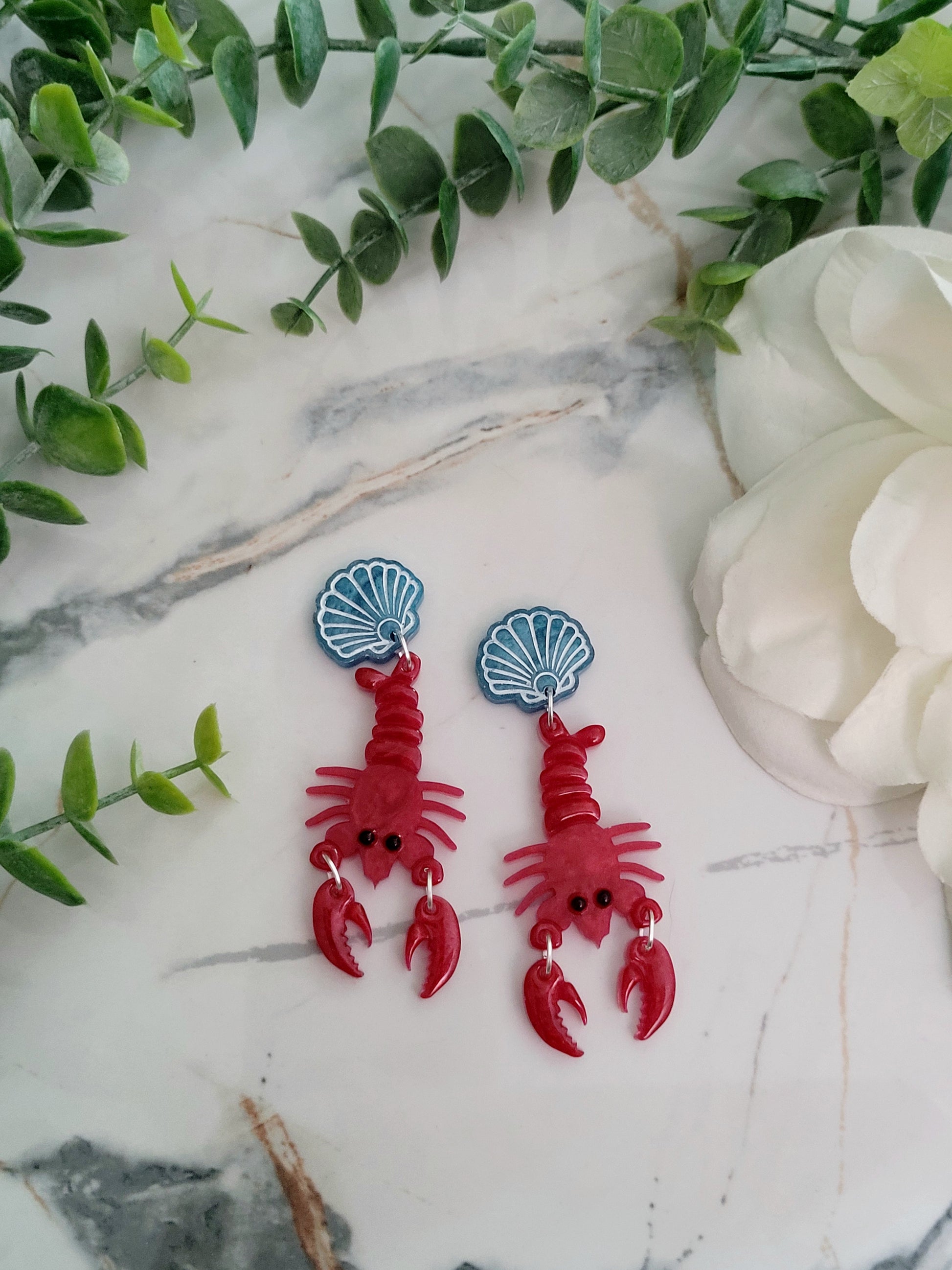 close up of Red Lobster with a blue shell stud earrings on a marble background with foliage.