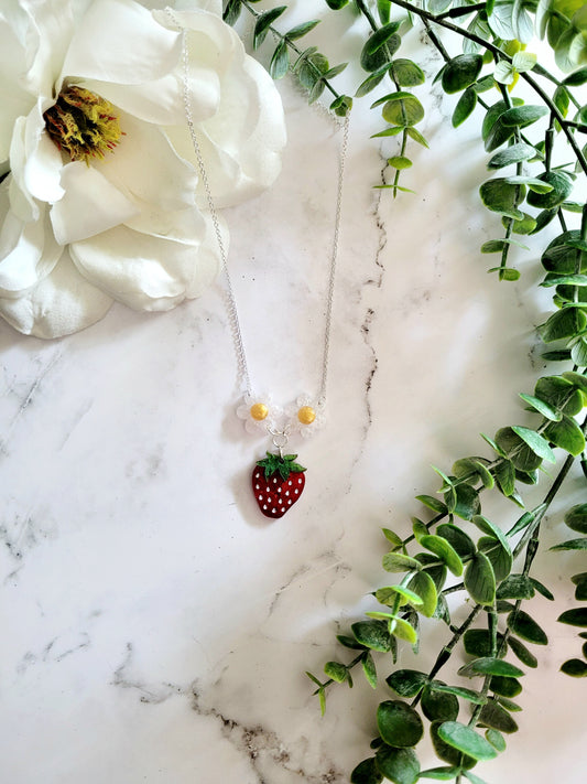 Resin Strawberry and flower necklace on a marble background surrounded by foliage.