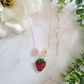 close up of Resin Strawberry and flower necklace on a marble background surrounded by foliage.