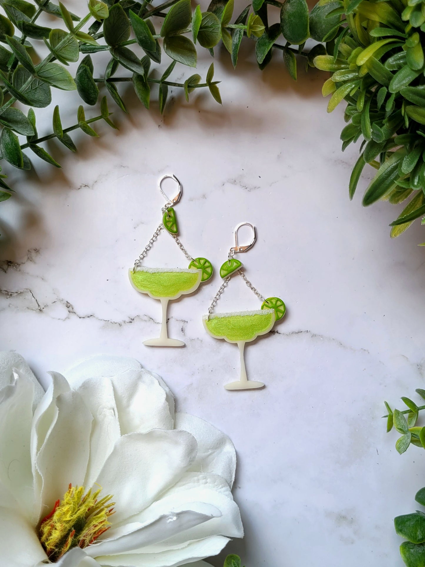 Lime margarita glass earrings on a marble background with foliage. 