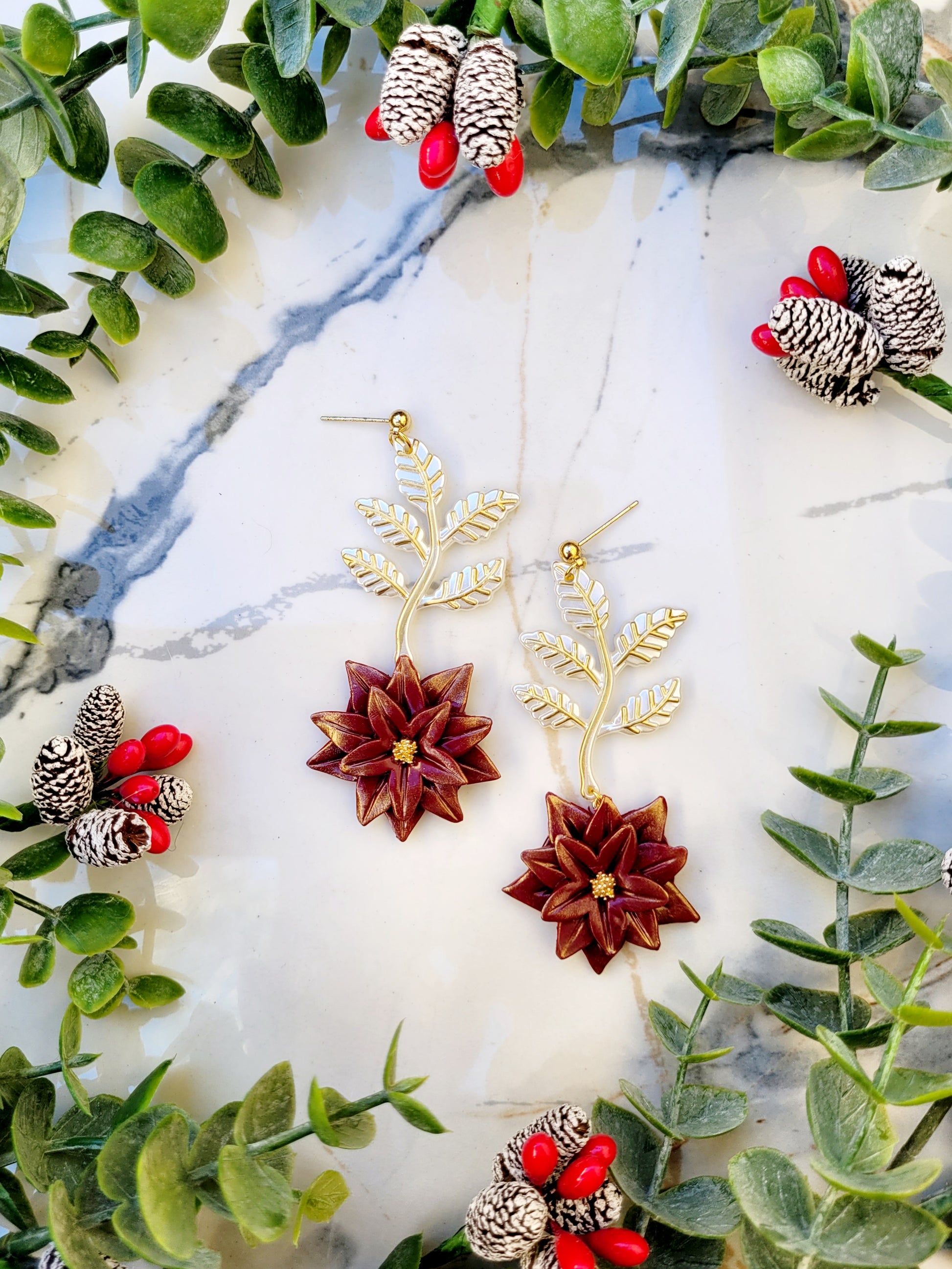 Red and gold poinsettia earrings on a marble background surrounded by foliage. 