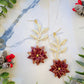 close up of Red and gold poinsettia earrings on a marble background surrounded by foliage. 