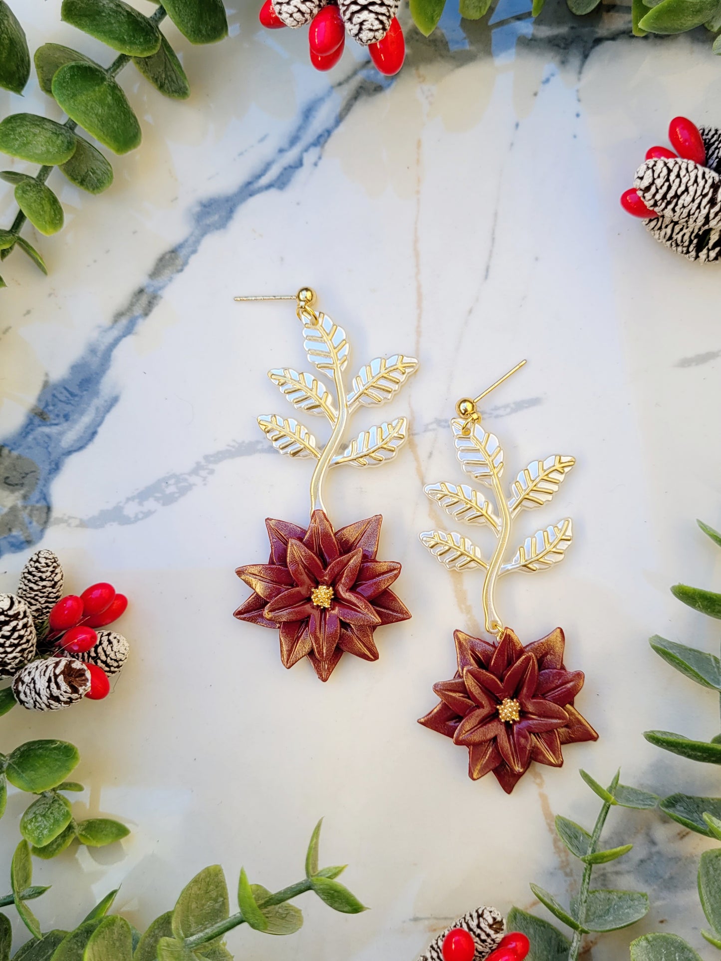 close up of Red and gold poinsettia earrings on a marble background surrounded by foliage. 