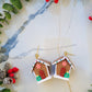 close up of stud Gingerbread house earrings on side on a marble background surrounded by foliage.
