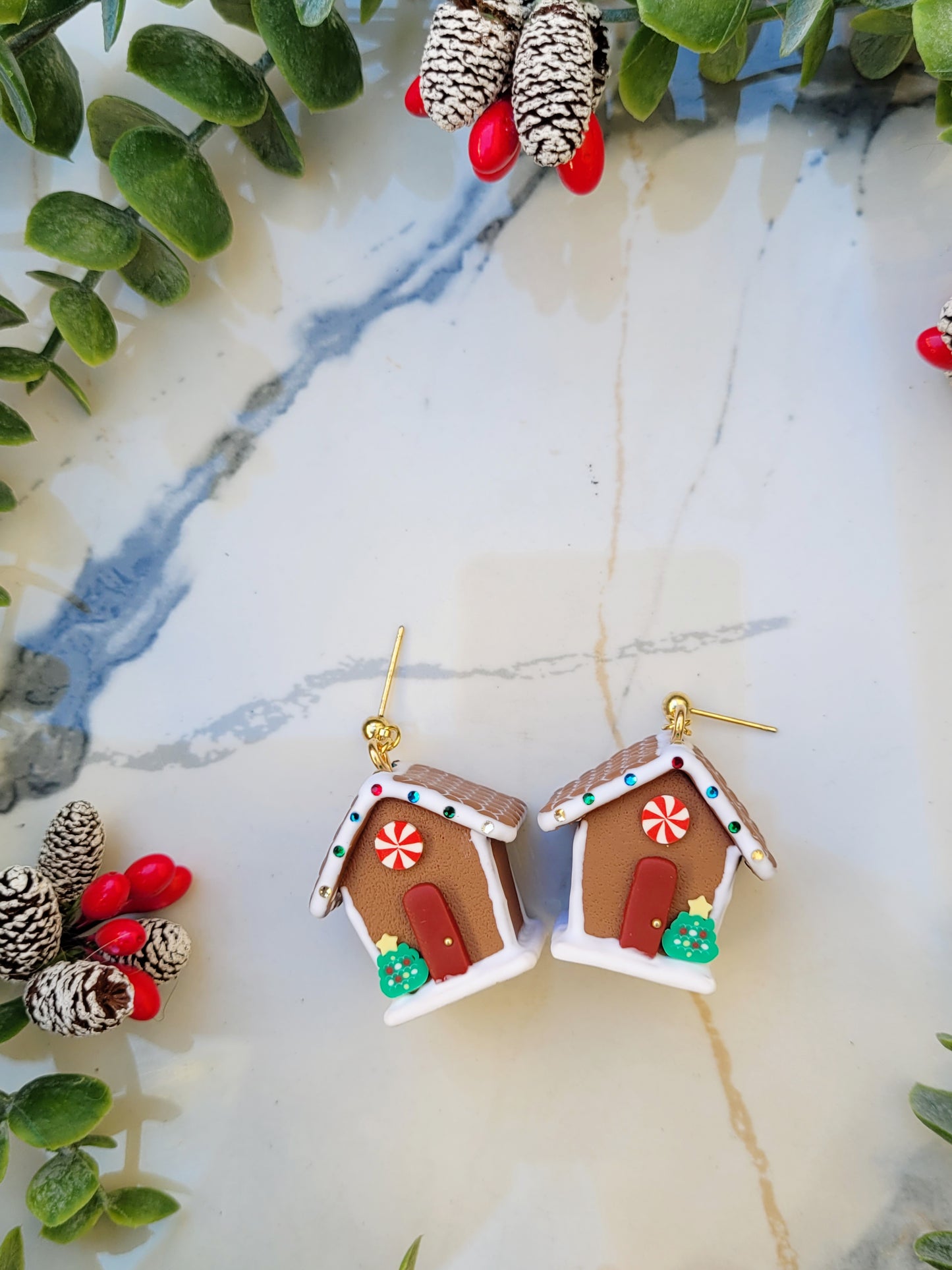 close up of stud Gingerbread house earrings on side on a marble background surrounded by foliage.